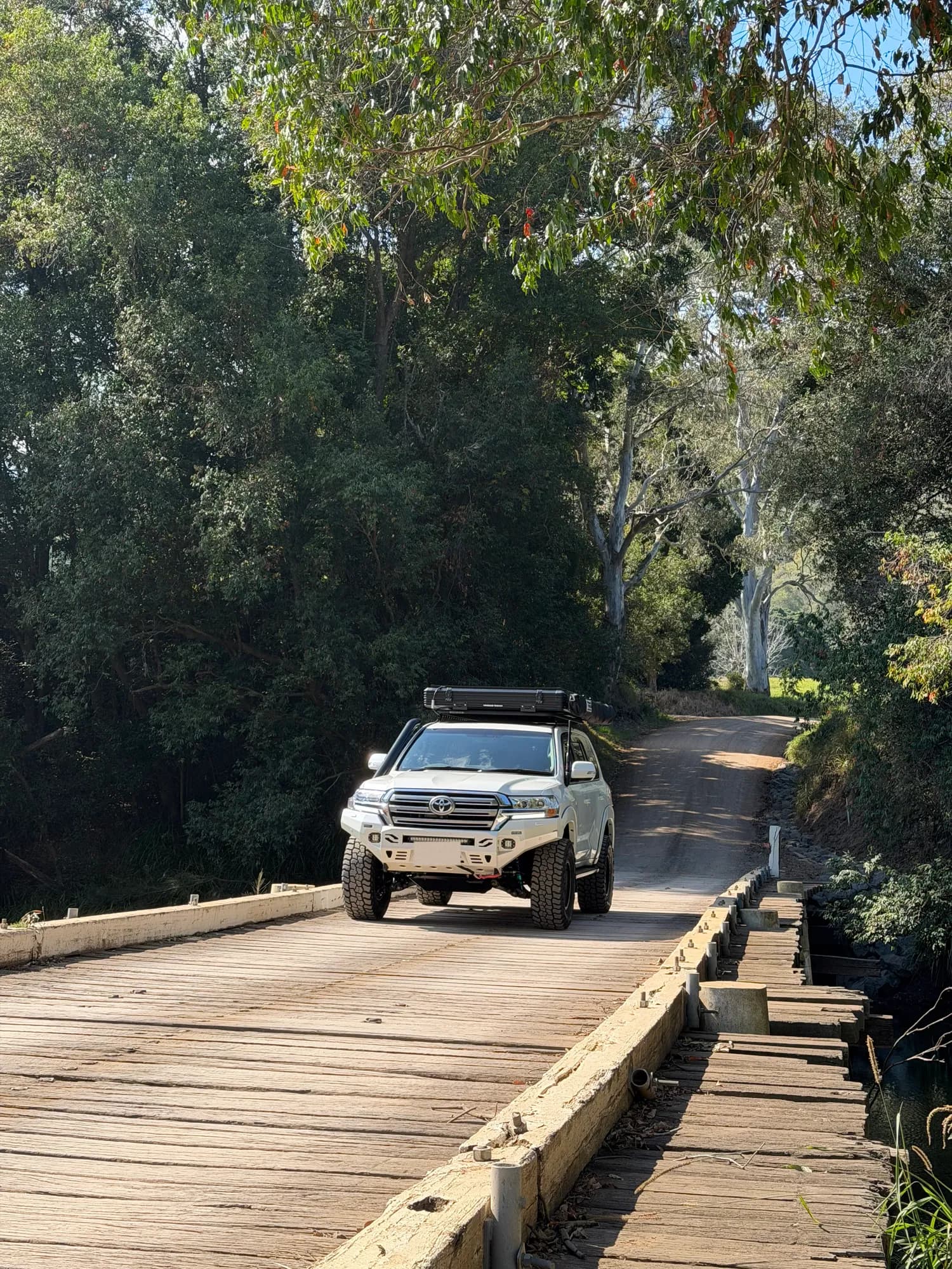 A white 200 Series with a rooftop tent driving across an old timber bridge under tall gum trees