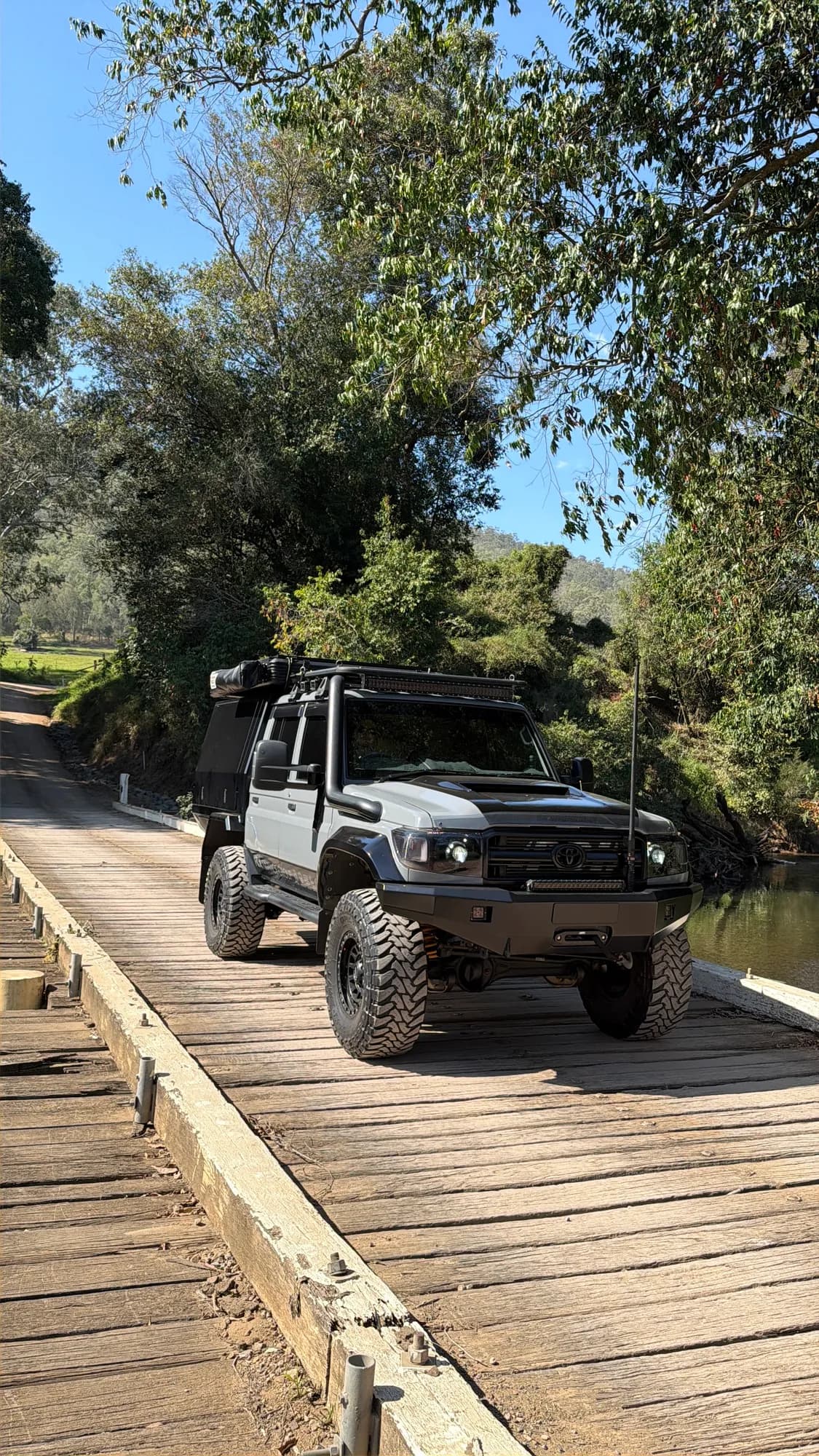 A grey 79 Series with a canopy and snorkel crossing an old timber bridge beside a creek