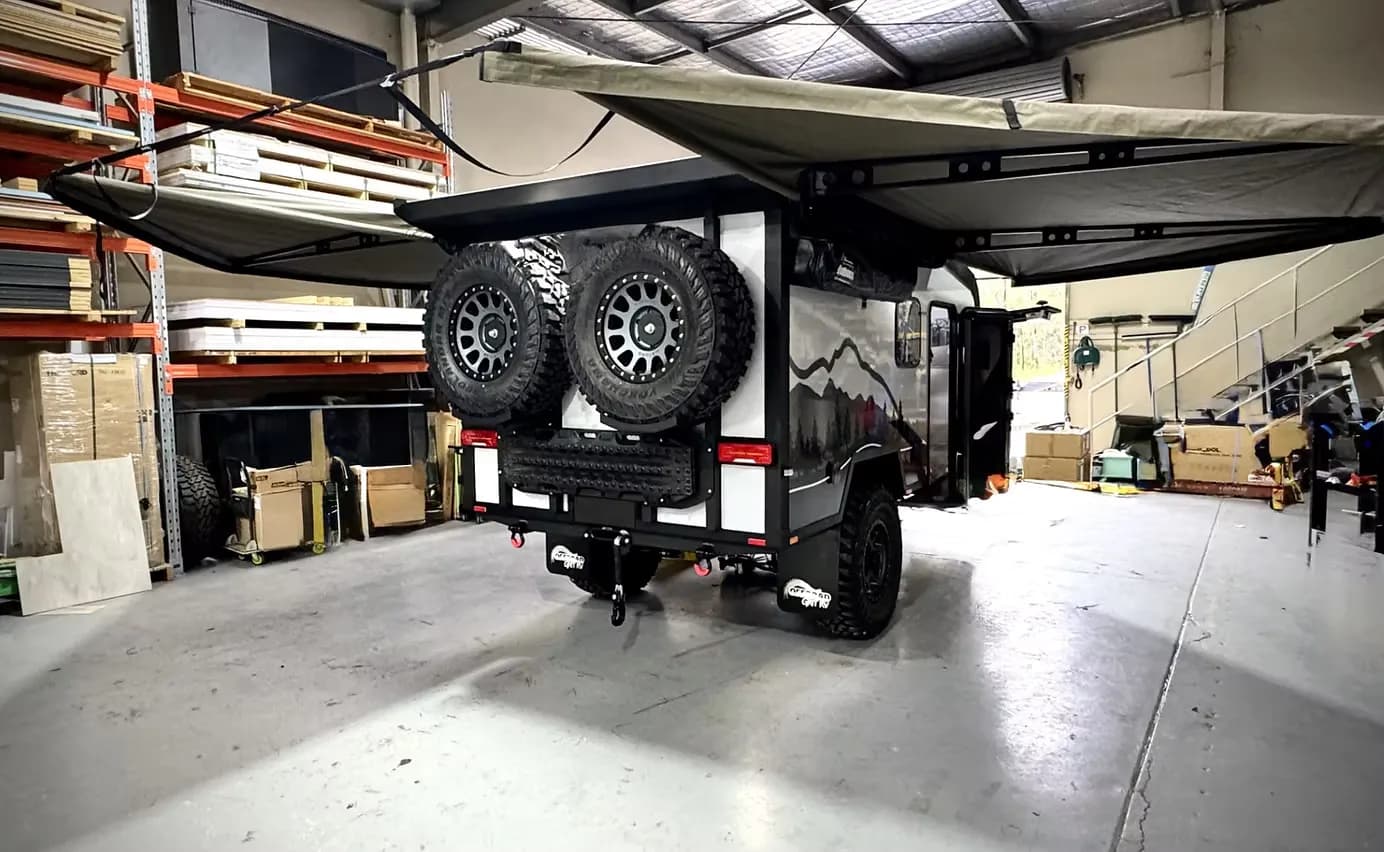 The rear of a compact off-road camper with twin spare wheels and its awning out, inside a build workshop