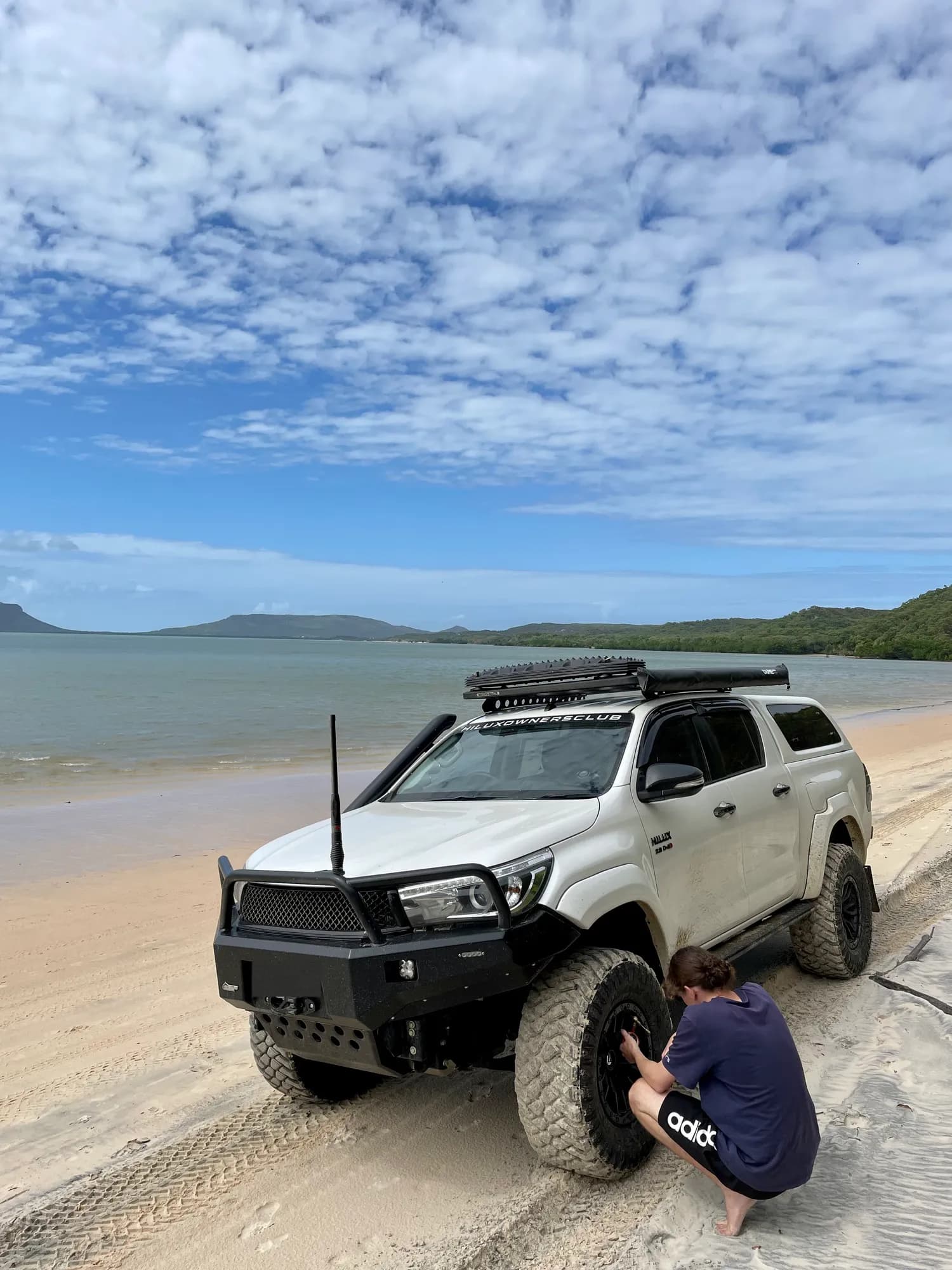 A white Hilux parked on a beach beside calm water while someone checks the tyre pressures