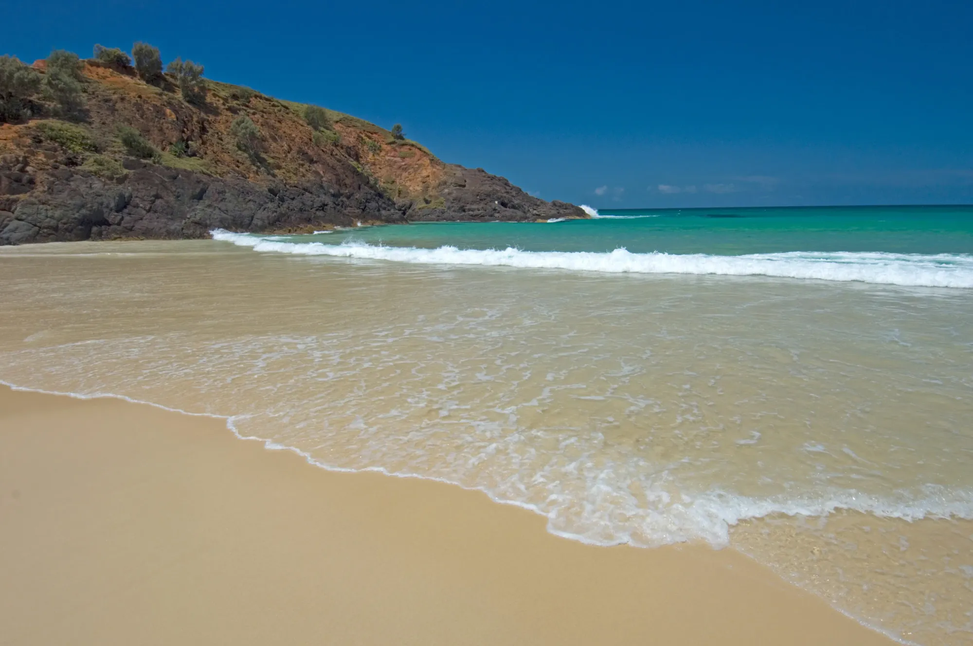 Clear turquoise water breaking on a white sand beach below a headland on Moreton Island