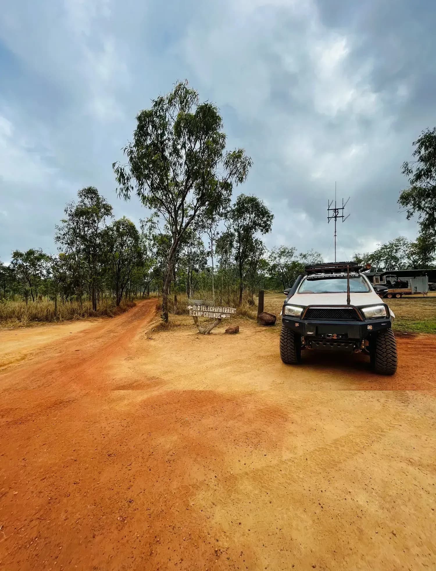A white Hilux parked on red dirt beside the Old Telegraph Track and camp grounds sign under a cloudy sky