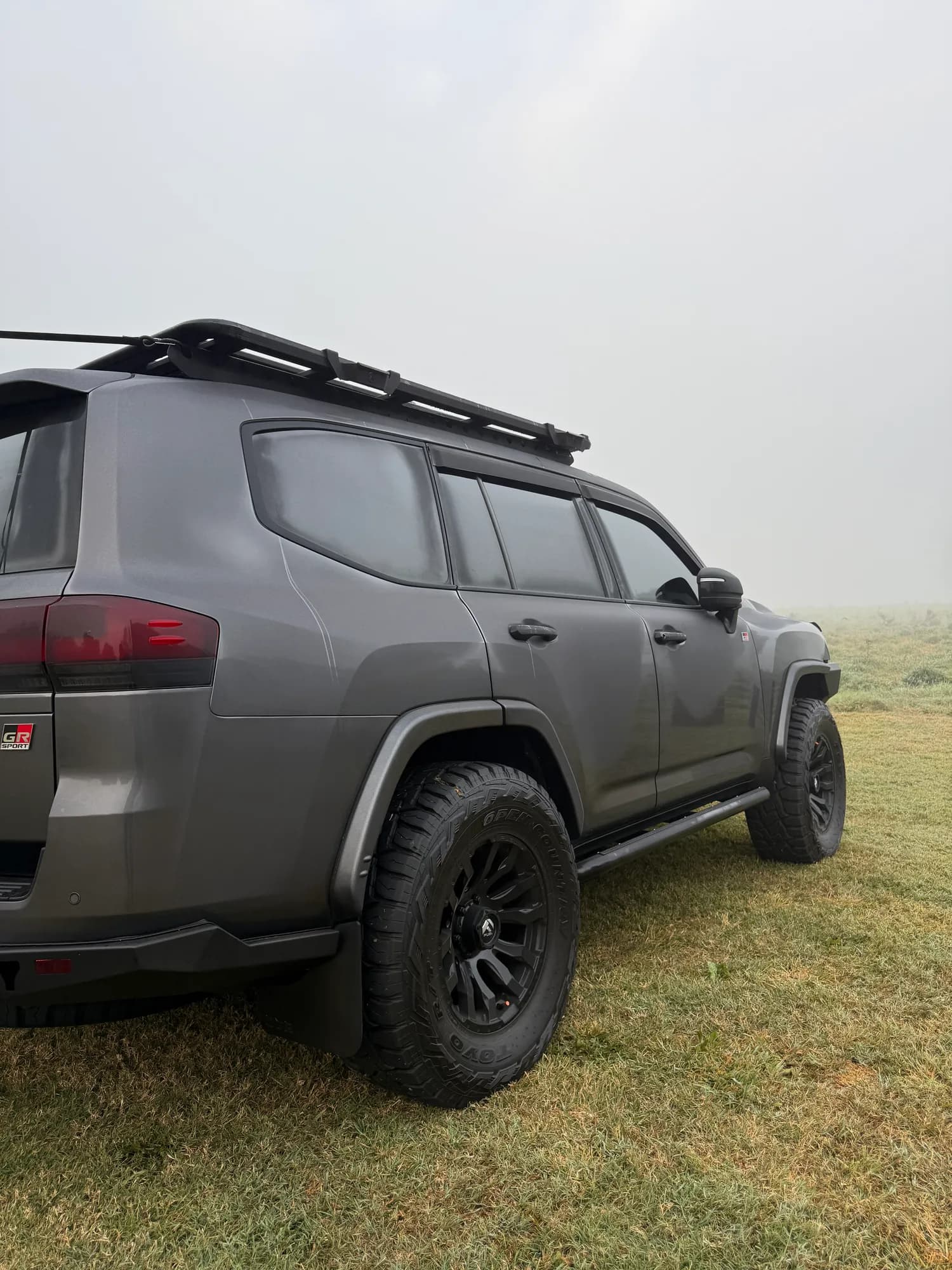 A grey 300 Series with a roof rack parked on wet grass in thick early morning fog, seen from the rear quarter